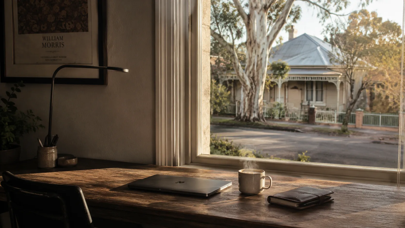 A wooden desk by a window in an Australian home study. Closed laptop, coffee mug, leather notebook on the desk. Through the window a Federation-style house and eucalyptus trees in late afternoon golden light.